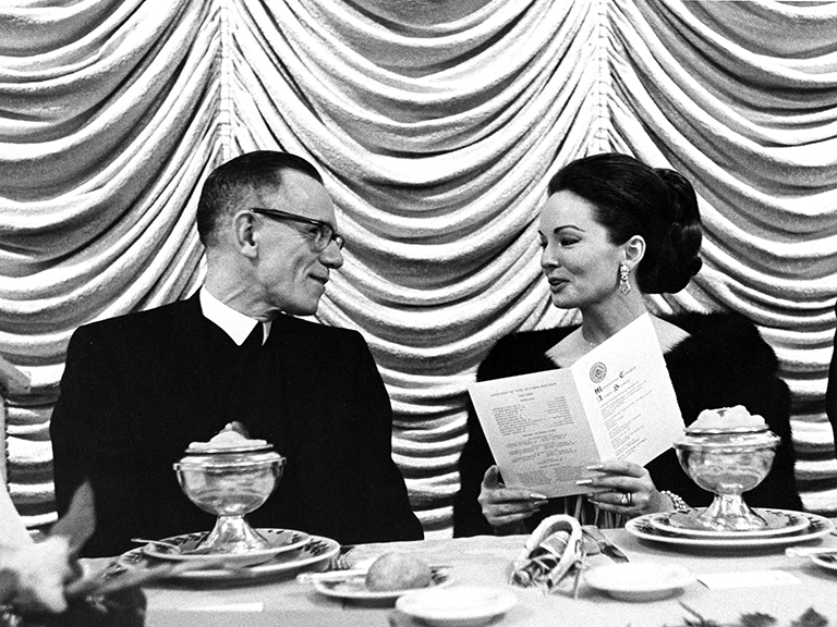 At the Manhattan College Alumni Banquet, actress Ann Blyth and College President Brother Gregory Nugent are seated at a large dais and are having a discussion.  Brother Gregory wears his religious garb and Ms. Blyth has a fancy fur shawl and she is holding the Alumni Banquet Program.  In front of them is the first course made up of fruit in fancy silver cups.